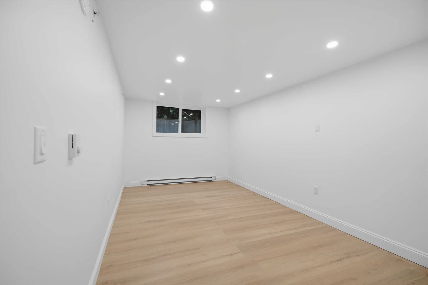 A bedroom photographed empty: bare floor, bare walls, window and baseboard heater.